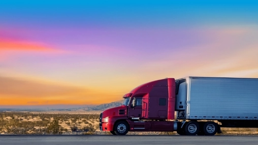 Red semi-truck with white trailer driving on a highway through a desert landscape under a vivid sunset sky