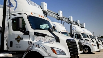Row of white zero-emissions electric semi-trucks at a depot with overhead charging equipment attached
